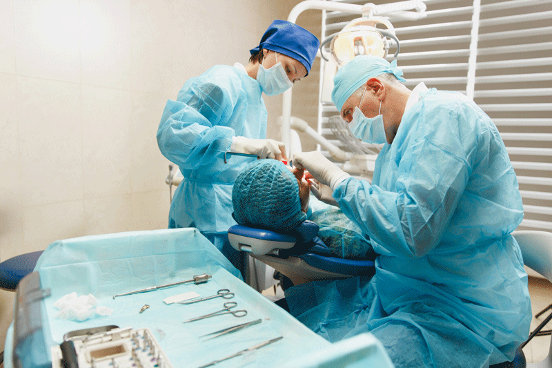Male old professional dentist surgeon in uniform and female assistant helps to performing operation install dental implant teeth of woman patient in clinic light office with modern tools equipment.